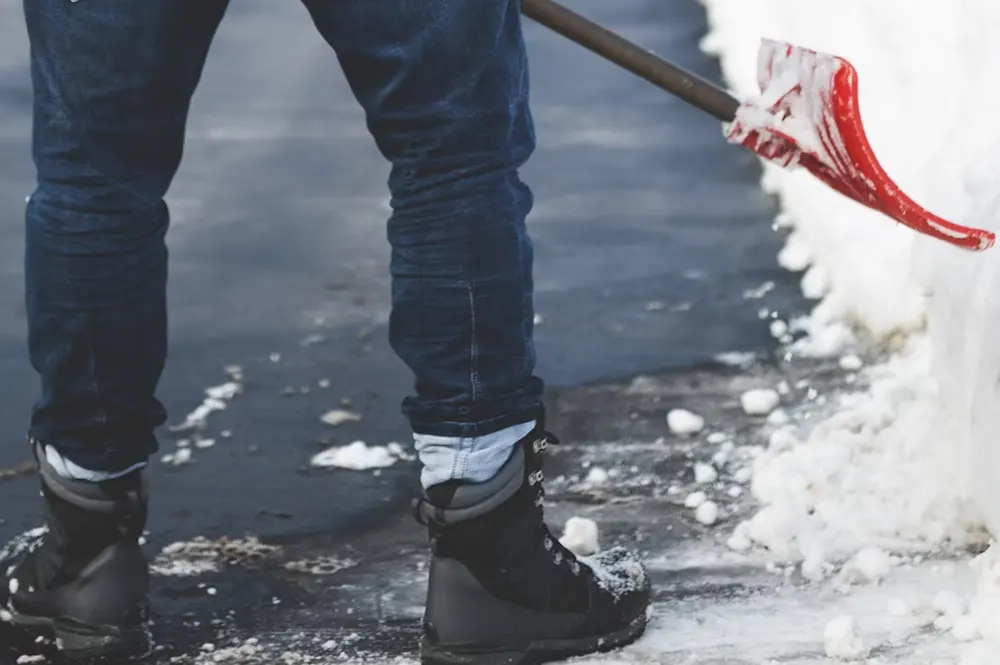 Person wearing jeans and black boots shoveling snow off pavement.